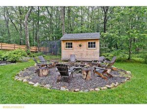 Rustically styled, six chairs surrounding fire pit on gravel circle in the middle of lawn