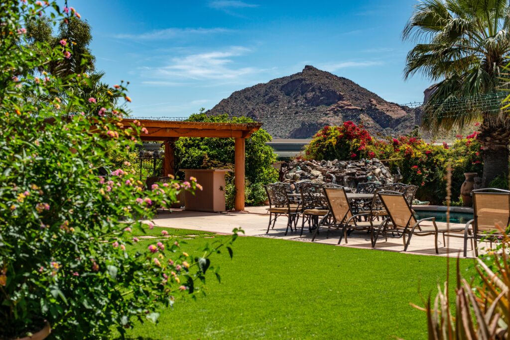 Outdoor patio on a sunny day, bush in the foreground, green grass, mountain in the background.